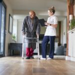 Senior Man In Dressing Gown Using Walking Frame Being Helped By Female Nurse With Clipboard